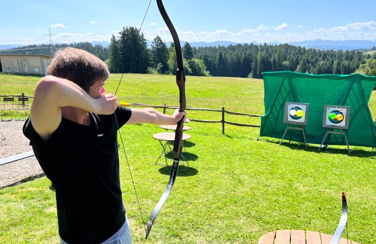 Ruhe finden und das Ziel im Blick behalten Eine Teilnehmerin konzentriert sich beim Bogenschießen auf einer Wiese vor Bergkulisse.