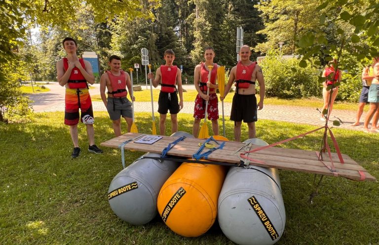 Gemeinsam gebaut – gemeinsam stolz Eine Gruppe steht mit Schwimmwesten und Paddeln hinter ihrem gemeinsam gebauten Floß.