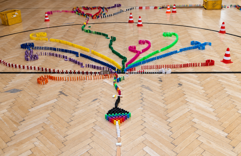 Domino Day – gemeinsam etwas schaffen Bunte Dominobahnen verbinden sich zu einer gemeinsamen Kettenreaktion auf einem Hallenboden.